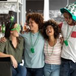 Four people with their arms around each other laughing and smiling. Each person is wearing St. Patrick's Day themed accessories.