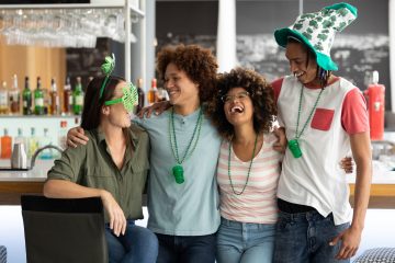 Four people with their arms around each other laughing and smiling. Each person is wearing St. Patrick's Day themed accessories.