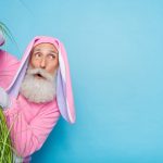 An old man with a white beard, dressed in a pink bunny costume, peeking from behind a plant against a bright blue background.